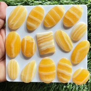Pode incluir: Uma cole&ccedil;&atilde;o de catorze caboch&otilde;es de pedra polida com faixas amarelas e brancas dispostos num suporte quadrado branco. As pedras apresentam superf&iacute;cies lisas e brilhantes com padr&otilde;es de camadas horizontais e verticais em tons de &acirc;mbar.