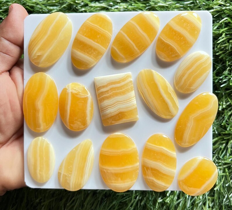 Pode incluir: Uma cole&ccedil;&atilde;o de catorze caboch&otilde;es de pedra polida com faixas amarelas e brancas dispostos num suporte quadrado branco. As pedras apresentam superf&iacute;cies lisas e brilhantes com padr&otilde;es de camadas horizontais e verticais em tons de &acirc;mbar.