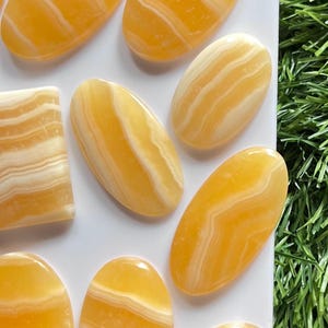 Pode incluir: Uma cole&ccedil;&atilde;o de caboch&otilde;es de pedra polida com faixas amarelas e brancas dispostos sobre uma superf&iacute;cie branca. As pedras s&atilde;o principalmente de formato oval com uma pe&ccedil;a retangular, cada uma exibindo superf&iacute;cies lisas e transl&uacute;cidas.