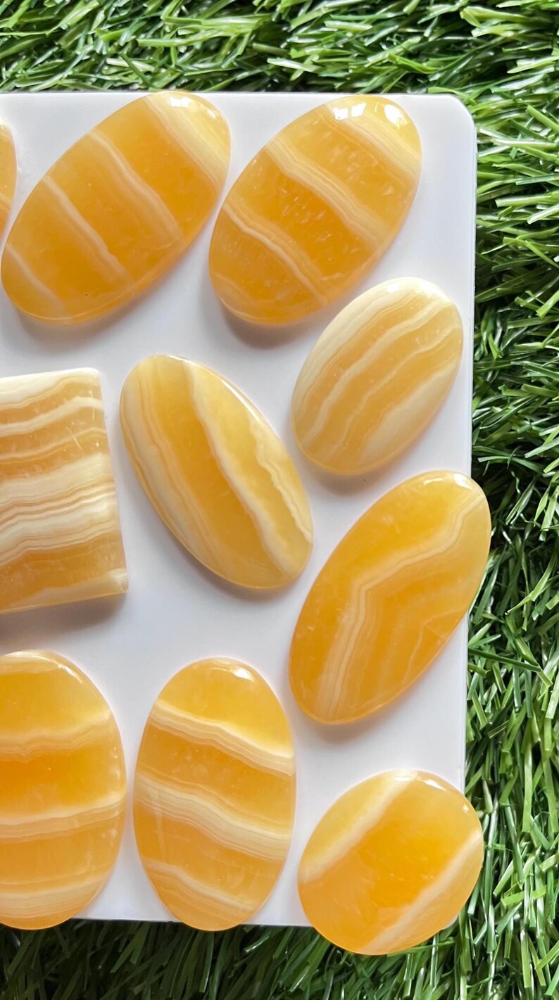 Pode incluir: Uma cole&ccedil;&atilde;o de caboch&otilde;es de pedra polida com faixas amarelas e brancas dispostos sobre uma superf&iacute;cie branca. As pedras s&atilde;o principalmente de formato oval com uma pe&ccedil;a retangular, cada uma exibindo superf&iacute;cies lisas e transl&uacute;cidas.