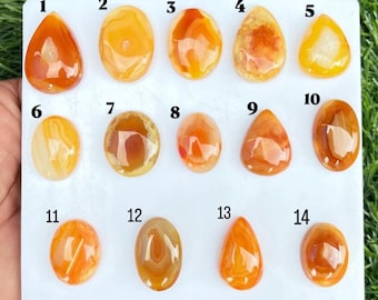 Pode incluir: Uma coleção de catorze pedras preciosas polidas e lisas, lapidadas em cabochão, em tons de laranja, amarelo e âmbar. As pedras estão dispostas num cartão de apresentação retangular branco, cada uma numerada de um a catorze com algarismos pretos.