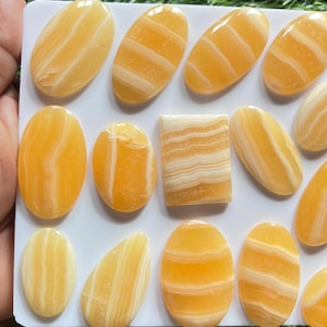 Pode incluir: Uma cole&ccedil;&atilde;o de caboch&otilde;es de pedra polida com faixas laranja e brancas dispostos numa placa retangular branca. As pedras apresentam v&aacute;rias formas, incluindo ovais, um ret&acirc;ngulo e uma gota, todas com superf&iacute;cies lisas e camadas naturais.