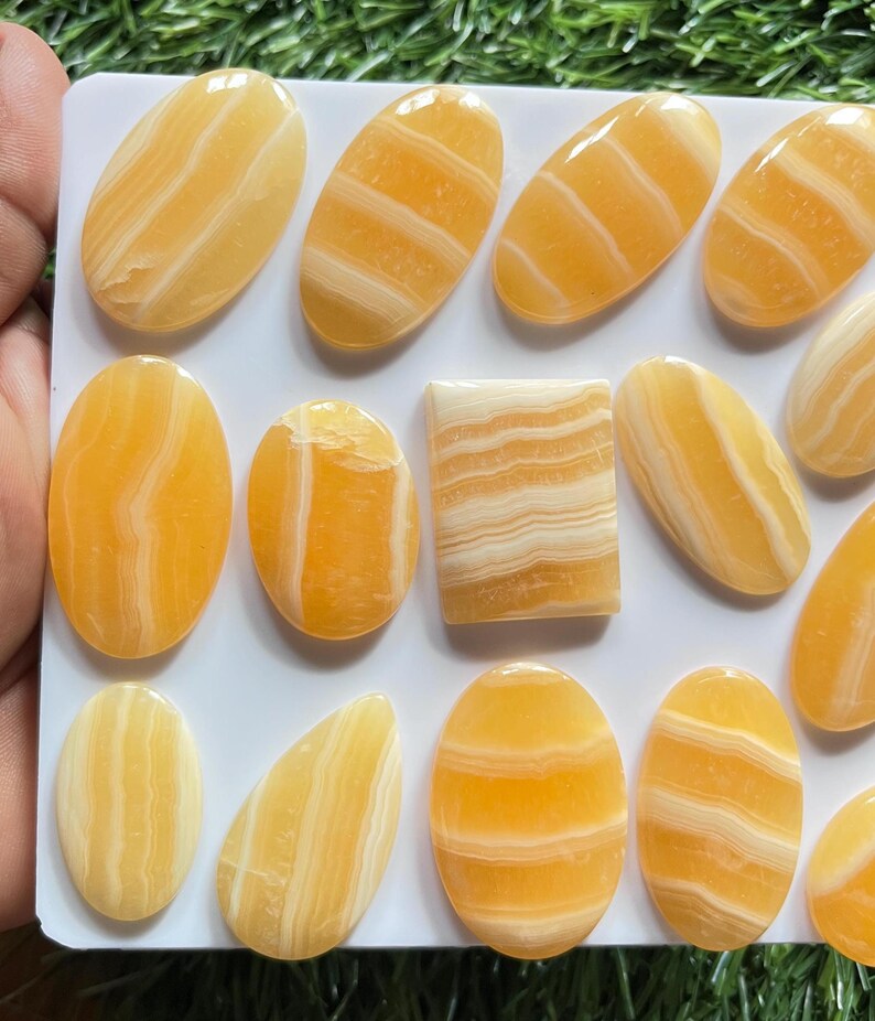 Pode incluir: Uma cole&ccedil;&atilde;o de caboch&otilde;es de pedra polida com faixas laranja e brancas dispostos numa placa retangular branca. As pedras apresentam v&aacute;rias formas, incluindo ovais, um ret&acirc;ngulo e uma gota, todas com superf&iacute;cies lisas e camadas naturais.