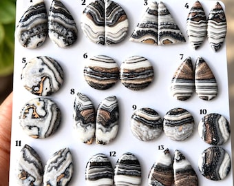 Puede incluir: Una colección de trece pares o cabujones individuales pulidos dispuestos sobre un tablero blanco. Cada piedra presenta patrones de bandas distintos en tonos blanco, negro, gris y cobre, numerados del uno al trece para su identificación.