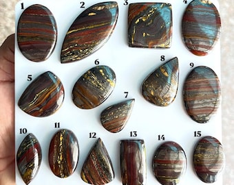 Puede incluir: Una colección de quince cabujones de hierro de tigre pulidos dispuestos en un tablero blanco, numerados del 1 al 15. Cada piedra preciosa presenta bandas horizontales y arremolinadas de color rojo intenso, oro metálico y capas de hematita gris.