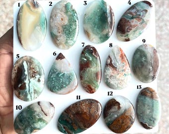 Puede incluir: Trece piedras preciosas cabujón pulidas, de forma ovalada y de lágrima, dispuestas sobre un tablero blanco. Cada piedra presenta patrones únicos de inclusiones verdes, marrones y crema. Numeradas del 1 al 13 en color negro.