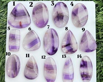 Puede incluir: Una colección de catorce piedras preciosas pulidas y lisas, talladas en cabujón, con patrones translúcidos de bandas blancas y moradas. Cada piedra está numerada del 1 al 14 y muestra zonas de color únicas e irregulares en formas variadas como óvalos y gotas.