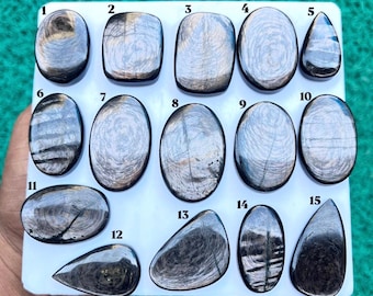 Puede incluir: Una colección de quince piedras preciosas cabujón pulidas, de color gris oscuro y aspecto metálico, dispuestas sobre un tablero blanco. Cada piedra presenta patrones únicos, arremolinados o lineales, y está numerada del uno al quince.
