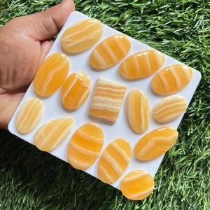 Pode incluir: Uma cole&ccedil;&atilde;o de quinze pedras de calcita polidas com faixas amarelas e brancas dispostas em um suporte quadrado branco. As pedras s&atilde;o principalmente de formato oval, com uma pe&ccedil;a retangular apresentando camadas horizontais de amarelo e branco.