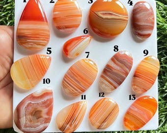 Pode incluir: Treze cabochões de ágata bandada, polidos e lisos, em tons de laranja e vermelho, dispostos num painel branco. Cada pedra está numerada de 1 a 13 e apresenta padrões naturais únicos de riscas horizontais ou concêntricas em tons de âmbar e ferrugem.