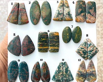 Puede incluir: Una colección de doce pares de cabujones de jaspe pulidos y multicolores dispuestos sobre un tablero blanco. Cada par está numerado del 1 al 12 y presenta patrones únicos de inclusiones minerales verdes, rojas, marrones y amarillas.
