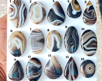 Puede incluir: Una colección de quince cabujones de ágata pulidos dispuestos sobre un tablero blanco, numerados del uno al quince. Cada piedra presenta patrones de bandas distintos en tonos marrón, blanco, gris y negro, con formas ovaladas y de lágrima.