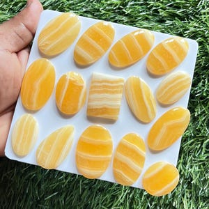 Pode incluir: Uma cole&ccedil;&atilde;o de catorze caboch&otilde;es de calcita polida com faixas amarelas e brancas dispostos numa placa quadrada branca. Treze pedras s&atilde;o de formato oval, enquanto uma pe&ccedil;a central &eacute; retangular com cantos arredondados e faixas horizontais.