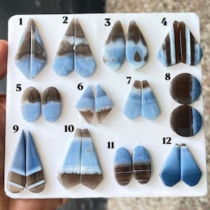 May include: A collection of twelve pairs of polished, smooth, blue and brown banded gemstones arranged on a white display board. Each pair is numbered from one to twelve, showcasing various geometric shapes including triangles, ovals, and circles.