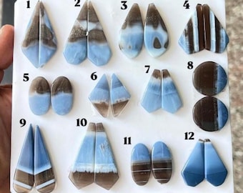 Pode incluir: Uma coleção de doze pares de pedras preciosas polidas, lisas, com faixas azuis e castanhas, dispostas num painel branco. Cada par está numerado de um a doze, apresentando várias formas geométricas, incluindo triângulos, ovais e círculos.