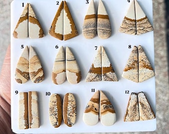 Puede incluir: Una colección de doce pares de cabujones de jaspe pulidos con reverso plano, dispuestos sobre un tablero blanco. Cada par presenta patrones naturales en tonos tierra, en matices beige, crema, marrón y negro, numerados del 1 al 12.