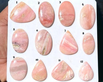 Puede incluir: Una colección de doce cabujones de ópalo rosa pulidos dispuestos en una tarjeta de presentación blanca, numerados del uno al doce. Cada gema presenta patrones únicos en tonos rosa suave, crema y blanco con un acabado liso y brillante.