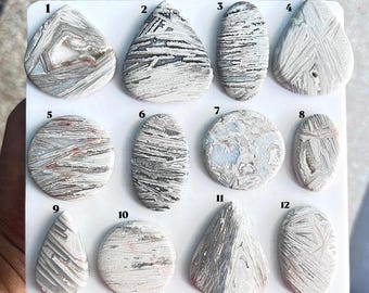 Puede incluir: Una colección de doce cabujones pulidos dispuestos sobre un tablero blanco, numerados del uno al doce. Estas piedras presentan patrones intrincados en gris, blanco y beige con formas variadas que incluyen gotas, círculos y óvalos.