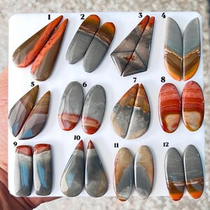 May include: A collection of twelve pairs of polished, multi-colored cabochon gemstones arranged on a white display board. Each pair is numbered from 1 to 12, featuring various shapes including teardrop, oval, and rectangular, with earthy tones.