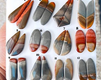 Pode incluir: Uma coleção de doze pares de pedras preciosas cabochon polidas e multicoloridas dispostas num painel branco. Cada par está numerado de 1 a 12, apresentando várias formas, incluindo gota, oval e retangular, com tons terrosos.
