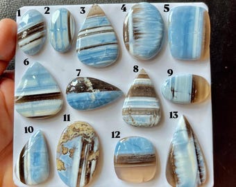 Puede incluir: Una colección de trece cabujones de ópalo pulidos con bandas azules y marrones dispuestos en un tablero blanco. Cada piedra preciosa presenta patrones de rayas horizontales o verticales únicos en tonos azul claro, blanco y marrón oscuro.