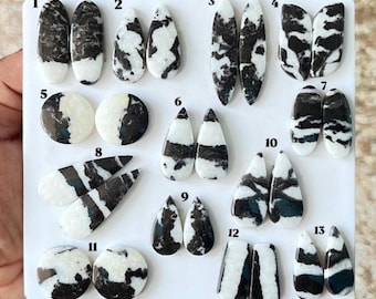 Puede incluir: Una colección de trece pares de cabujones pulidos con patrones en blanco y negro dispuestos sobre un tablero blanco. Cada par está numerado del uno al trece, presentando formas variadas que incluyen óvalos, gotas, círculos y rectángulos.