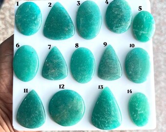 Puede incluir: Una colección de catorce cabujones de amazonita de color verde azulado, pulidos y dispuestos sobre un tablero blanco. Cada piedra está numerada del 1 al 14 y presenta formas variadas, incluyendo óvalos, gotas y círculos.
