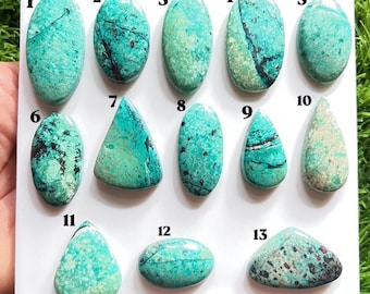 Puede incluir: Trece cabujones pulidos de color turquesa dispuestos sobre una tarjeta de presentación blanca, numerados del uno al trece. Cada piedra presenta patrones de matriz naturales únicos en negro, marrón y blanco, con formas variadas que incluyen óvalos, gotas y triángulos.