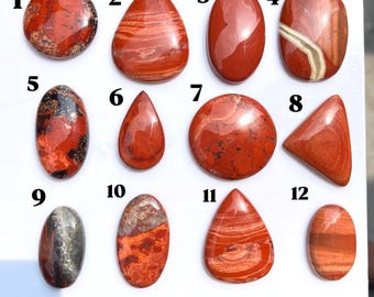 Puede incluir: Una colección de doce cabujones de jaspe rojo pulidos dispuestos en una cuadrícula sobre una superficie blanca. Cada piedra presenta patrones naturales únicos, incluyendo remolinos, bandas y motas en tonos rojos, naranjas y negros, numerados del uno al doce.