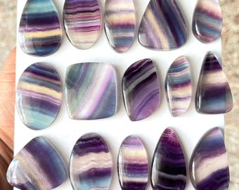 Puede incluir: Una colección de catorce cabujones de fluorita pulidos dispuestos en tres filas sobre una tarjeta de presentación blanca. Cada piedra preciosa presenta patrones de bandas distintos en tonos morado, azul, amarillo y blanco, con formas variadas como gotas, óvalos y rectángulos.