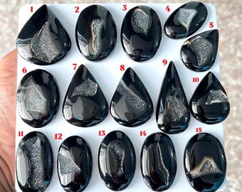 Puede incluir: Quince cabujones de ágata negra pulida dispuestos en una cuadrícula, cada uno con una inclusión central de cuarzo druzy. Las piedras están numeradas del uno al quince y varían en formas, incluyendo gotas, óvalos y formas redondeadas.