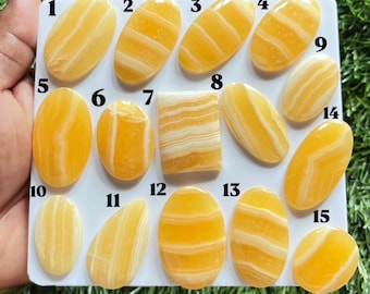 Puede incluir: Una colección de quince cabujones de ágata con bandas amarillas y blancas dispuestos en una tarjeta de presentación blanca. Las piedras están numeradas del 1 al 15 y presentan varias formas ovaladas, rectangulares y de lágrima con superficies lisas.