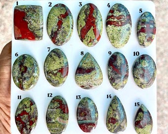 Puede incluir: Una colección de quince piedras preciosas pulidas en cabujón dispuestas sobre un tablero blanco, numeradas del 1 al 15. Cada piedra presenta un patrón único de inclusiones minerales de color verde oliva, rojo oscuro y gris.