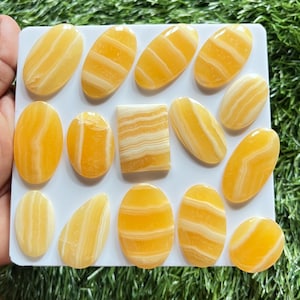 Pode incluir: Uma cole&ccedil;&atilde;o de catorze caboch&otilde;es de calcite amarela polida dispostos num expositor retangular branco. As pedras apresentam formas ovais e de gota suaves, com uma pe&ccedil;a retangular, todas exibindo faixas brancas distintas.