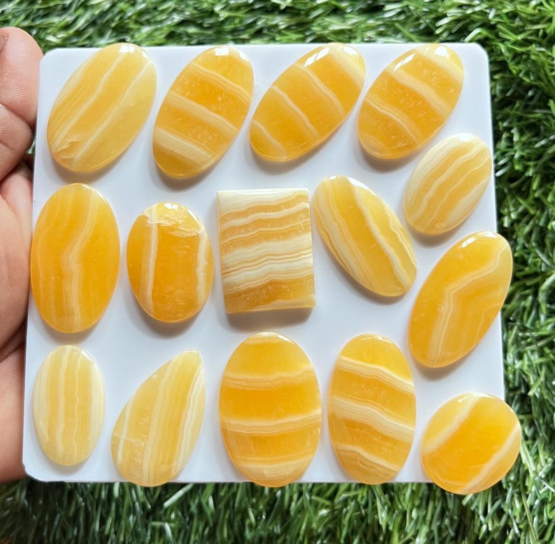 Pode incluir: Uma cole&ccedil;&atilde;o de catorze caboch&otilde;es de calcite amarela polida dispostos num expositor retangular branco. As pedras apresentam formas ovais e de gota suaves, com uma pe&ccedil;a retangular, todas exibindo faixas brancas distintas.