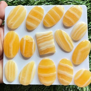 Pode incluir: Uma cole&ccedil;&atilde;o de catorze caboch&otilde;es de calcite polida com faixas amarelas e brancas, dispostos numa placa quadrada branca. As pedras apresentam formas lisas, ovais e retangulares com camadas minerais horizontais e verticais distintas.