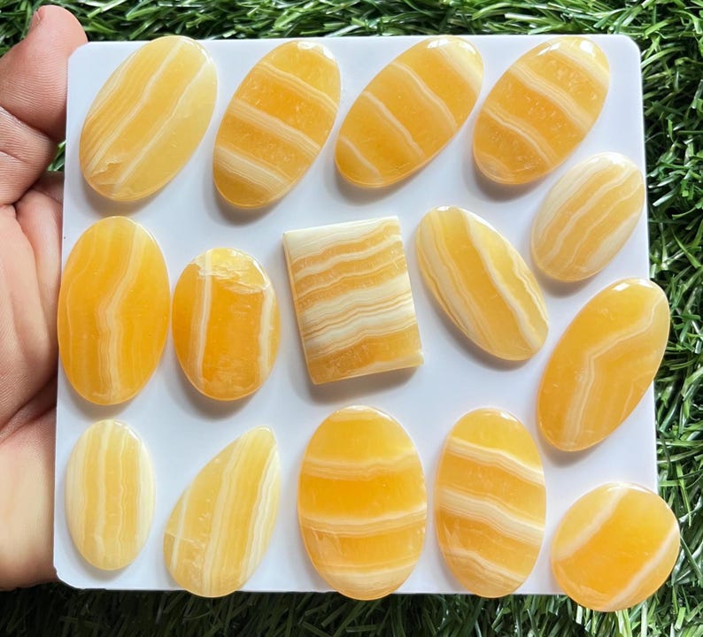 Pode incluir: Uma cole&ccedil;&atilde;o de catorze caboch&otilde;es de calcite polida com faixas amarelas e brancas, dispostos numa placa quadrada branca. As pedras apresentam formas lisas, ovais e retangulares com camadas minerais horizontais e verticais distintas.