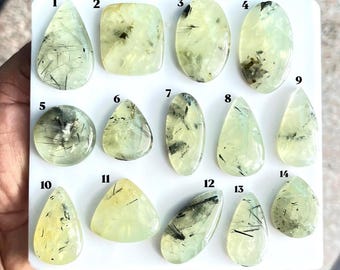 Puede incluir: Una colección de catorce piedras preciosas pulidas de color verde pálido con inclusiones negras en forma de aguja, dispuestas sobre un tablero blanco. Cada piedra está numerada del 1 al 14 y presenta formas únicas como gotas.