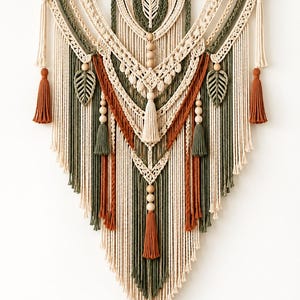 May include: A large macrame wall hanging suspended from a dark wooden branch. The design features cream, olive green, and terracotta cotton cords arranged in intricate knots, braided patterns, leaf-shaped accents, wooden beads, and hanging tassels.