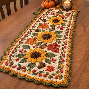 Pode incluir: Um caminho de mesa retangular de crochê com base creme, decorado com um padrão de girassóis amarelos, folhas de bordo laranja e vermelhas, folhas verdes, bolotas castanhas e pequenas bagas vermelhas, com uma borda verde festonada.