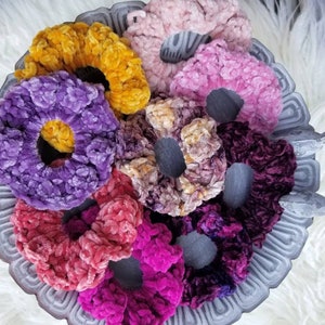 May include: A collection of colorful, crocheted hair scrunchies in various shades of purple, pink, yellow, and brown. The scrunchies are arranged on a gray, decorative plate.