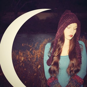May include: A woman wearing a long, burgundy knitted hat with a hood and matching fingerless gloves. The hat has long, dangling ties. She is standing in front of a crescent moon.