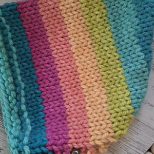 May include: A handmade, rainbow-striped knitted hat with a tag. The hat features a vibrant gradient of colors, including teal, pink, orange, and yellow. The hat is made of thick yarn and has long, braided ties.