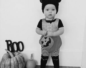 Puede incluir: Un niño vestido con un atuendo a rayas blanco y negro con un gorro de oso negro y un cubo de calabaza. El niño está de pie frente a una pared blanca con un letrero negro "BOO" y dos calabazas.