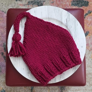 May include: A knitted burgundy hat with a tassel and a ball detail, resting on a white plate. The hat is made of thick yarn and has a ribbed brim. The tassel hangs down from the top of the hat. The plate is on a burgundy ottoman.