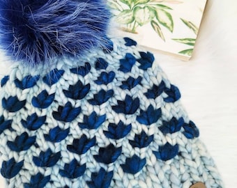 May include: A blue and white knitted beanie with a blue faux fur pom pom. The beanie has a pattern of blue flowers and a brown leather tag that says "Golden Hands Designs".