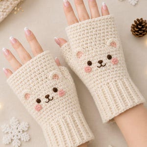 May include: A pair of cream-colored fingerless gloves made of knit yarn. Each glove features a bear face design with two small ears, dark brown eyes, a brown nose, and pink cheeks. The gloves have a ribbed cuff at the wrist and an open top.