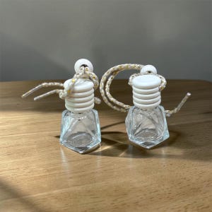 May include: Two small, clear glass bottles with a faceted, diamond-like shape. Each bottle features a white, ribbed wooden cap and a thick, beige cord with subtle gold metallic flecks threaded through the top for hanging purposes.