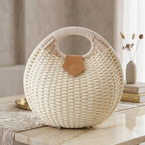 May include: A round, cream-colored woven straw handbag featuring a circular integrated handle and a tan leather-like triangular tab closure at the top center. The bag has a textured, basket-weave pattern and small feet at the base for stability.