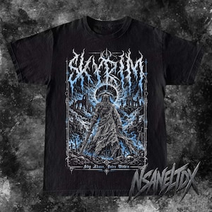 Op de afbeelding: Een zwart t-shirt met korte mouwen en een grafische print van een figuur in harnas met een kap, staand tussen ruïnes. De tekst SKYRIM staat in een gestileerd blauw en wit lettertype bovenaan, met de tekst Sky Above, Voice Within onderaan.